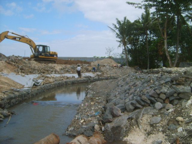 Excavator and workers building a rock gabion wall along the Keramas river bank to protect the surf break during early construction
